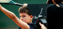 Child holding a tennis racket with a camera operator in the foreground on a tennis court.