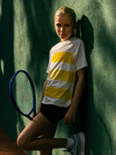 girl wearing a white and yellow striped shirt with a tennis racket against a green wall ROggio tennis apparel